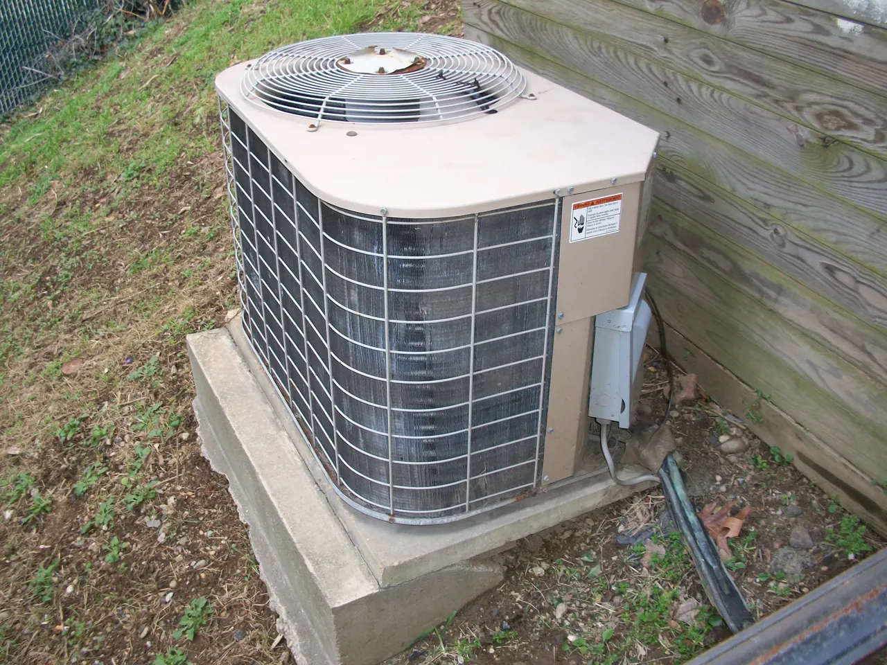 Older outdoor AC condenser on a concrete pad by a wooden fence